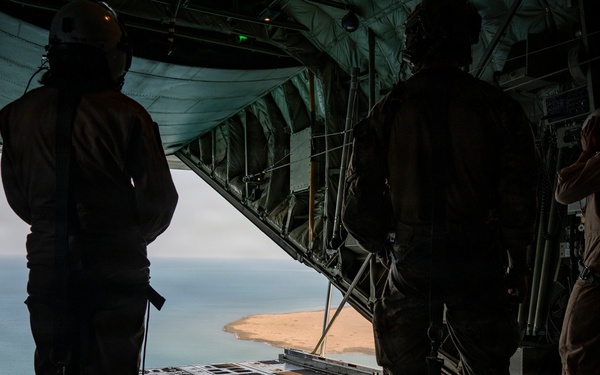 Three personnel are standing inside the open rear cargo ramp of a military transport aircraft, likely a C-130 Hercules, looking out over a coastal landscape below. The aircraft is in flight, with a sandy shoreline and body of water visible through the open ramp. The interior shows the characteristic roller-track cargo floor and structural elements of a military airlifter.