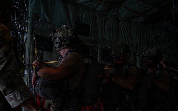 A group of military personnel in full tactical gear are lined up inside what appears to be a military transport aircraft, likely a C-130 or similar cargo plane. The soldiers are equipped with helmets, body armor, parachute packs, and tactical equipment, suggesting preparation for a airborne jump operation. The interior of the aircraft is dimly lit with a red cargo net/pallet visible in the background.