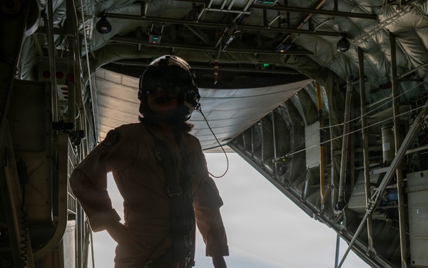 A military aircrew member stands silhouetted at the open rear cargo ramp of a military transport aircraft, likely a C-130 Hercules, in flight. The person is wearing a flight helmet with communications equipment and appears to be monitoring or observing from the open ramp. The interior of the cargo bay is visible with its characteristic ribbed structure, tie-down rings, and roller conveyor system.