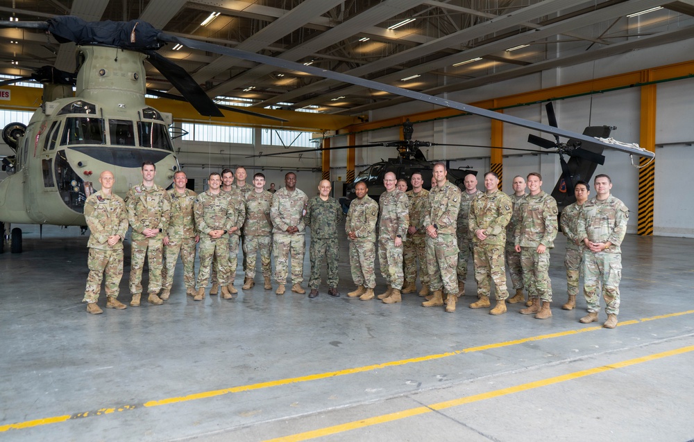 A group of approximately 18-20 military personnel in camouflage uniforms pose for a formal group photograph inside a military aviation hangar. Two military helicopters are visible in the background — a large CH-47 Chinook tandem-rotor helicopter on the left and what appears to be a UH-60 Black Hawk or AH-64 Apache on the right. The setting appears to be an official visit or unit photo opportunity.