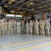 A group of approximately 18-20 military personnel in camouflage uniforms pose for a formal group photograph inside a military aviation hangar. Two military helicopters are visible in the background — a large CH-47 Chinook tandem-rotor helicopter on the left and what appears to be a UH-60 Black Hawk or AH-64 Apache on the right. The setting appears to be an official visit or unit photo opportunity.