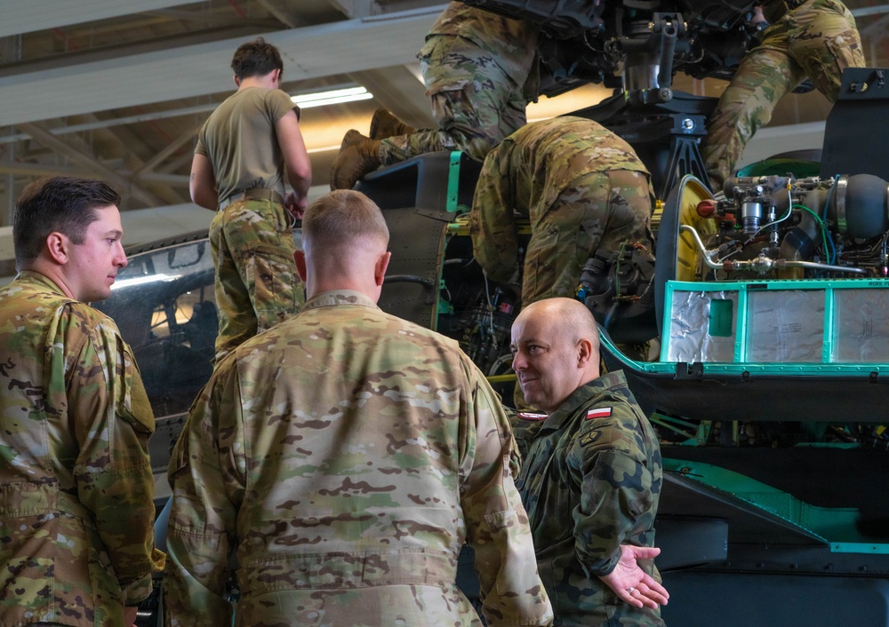 Military personnel in a hangar are performing maintenance or inspection on what appears to be a helicopter. A mix of soldiers wearing US Army OCP (Operational Camouflage Pattern) uniforms and at least one soldier wearing a Polish military uniform with a visible Polish flag patch are gathered around the aircraft. Some personnel are working on top of or inside the aircraft while others appear to be in discussion.