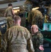 Military personnel in a hangar are performing maintenance or inspection on what appears to be a helicopter. A mix of soldiers wearing US Army OCP (Operational Camouflage Pattern) uniforms and at least one soldier wearing a Polish military uniform with a visible Polish flag patch are gathered around the aircraft. Some personnel are working on top of or inside the aircraft while others appear to be in discussion.