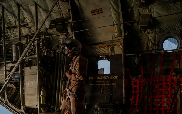 A military aircrew member stands near the open rear ramp of a large military transport helicopter or aircraft, likely a CH-47 Chinook, while in flight over water. The crew member is wearing flight gear including a helmet with communications equipment and a tan flight suit. The interior shows standard military aircraft features including an emergency exit door, red cargo netting, and a lowered loading ramp.