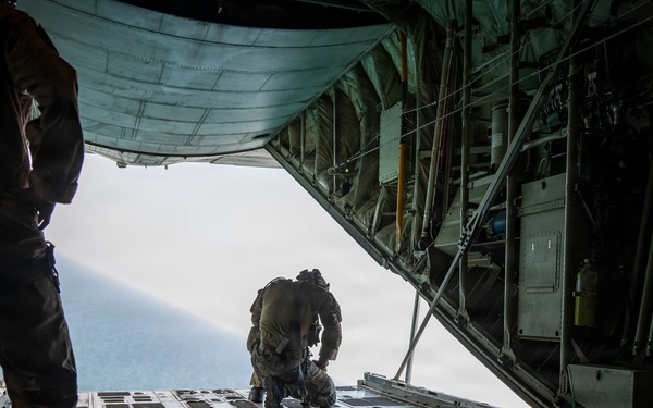 The image shows the interior of a military cargo aircraft, likely a C-130 Hercules, with its rear ramp open in flight. Three military personnel are visible, with one crouching near the edge of the open ramp, apparently preparing for or in the process of a parachute jump. The view through the open ramp shows sky and what appears to be ocean or water below.