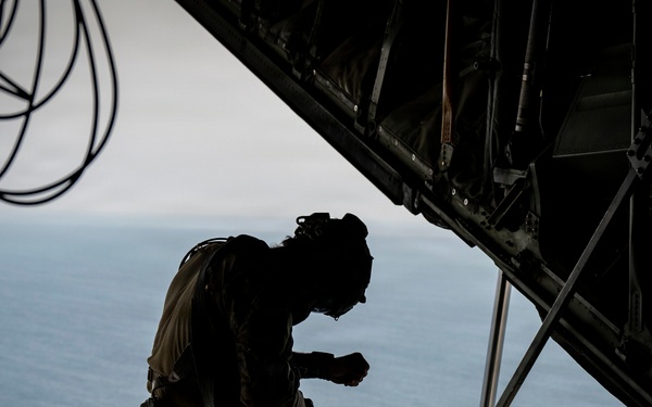 A military personnel member is kneeling on the open rear ramp of what appears to be a military transport aircraft, likely a C-130 Hercules, in flight over a large body of water. The figure is silhouetted against the open sky and ocean below, wearing full tactical gear and a helmet. The dramatic composition highlights the open ramp environment and the vast expanse of water visible beneath the aircraft.