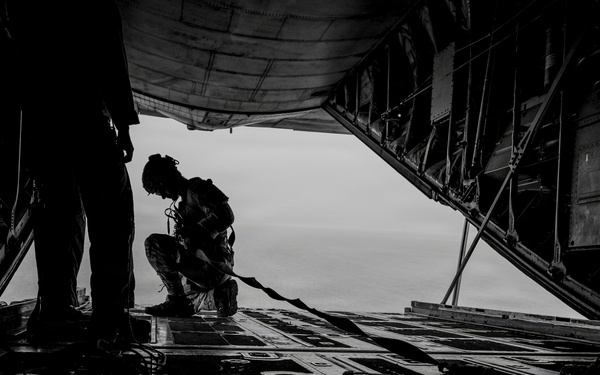 A black and white photograph taken from inside a military cargo aircraft, showing two silhouetted figures near the open rear ramp. One person is standing while another is crouching or kneeling near the edge of the open ramp, with sky visible in the background. The dramatic low-angle perspective emphasizes the aircraft's cargo floor and the open ramp opening.