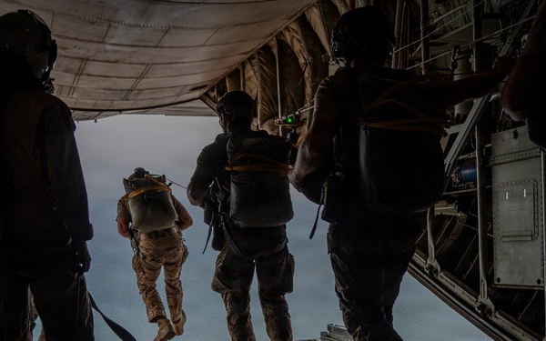 Military personnel are conducting a parachute jump operation from the open rear ramp of a large military cargo aircraft, likely a C-130 Hercules or similar transport. One jumper is already airborne and exiting the aircraft while others stand in queue on the ramp awaiting their turn. The scene is captured in dramatic silhouette against an overcast sky above what appears to be a body of water.