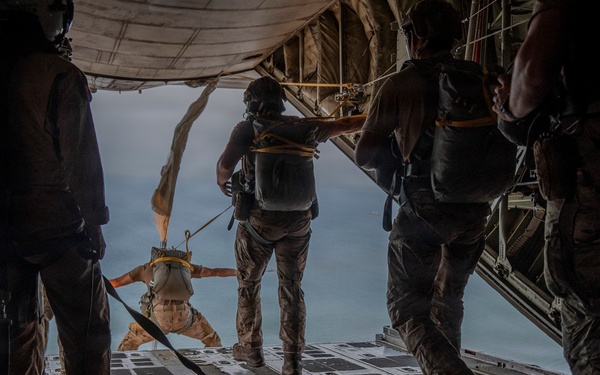 Military personnel are conducting a static-line parachute jump from the open rear cargo ramp of a military transport aircraft, likely a C-130 Hercules. One jumper is already exiting the aircraft in a horizontal position, while several others stand on the ramp observing or preparing to jump. The scene is set over open water or coastal terrain visible in the background.
