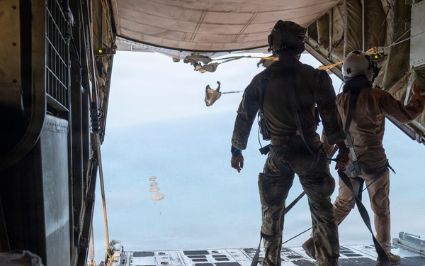 Two military personnel in combat gear stand at the open rear ramp of a large military transport aircraft, likely a C-130 Hercules, observing parachutes visible in the distance below. The scene depicts an active airborne operation with personnel or cargo already deployed. The image is taken from inside the aircraft looking outward toward the sky and terrain below.