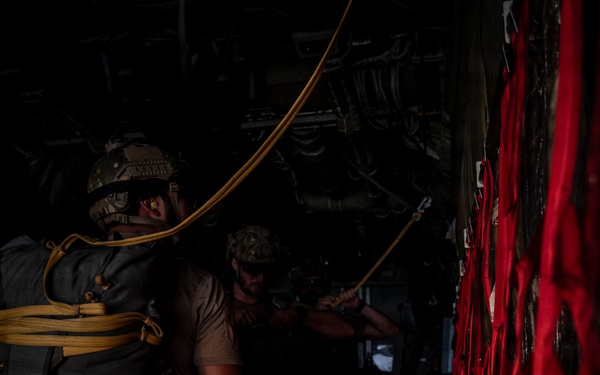 Two military personnel are visible inside what appears to be the cargo hold of a military aircraft, likely preparing for a parachute jump operation. The foreground figure is wearing a tactical helmet and carrying a large pack secured with yellow static line straps. A red cargo net or safety netting is visible along the right side of the aircraft interior.