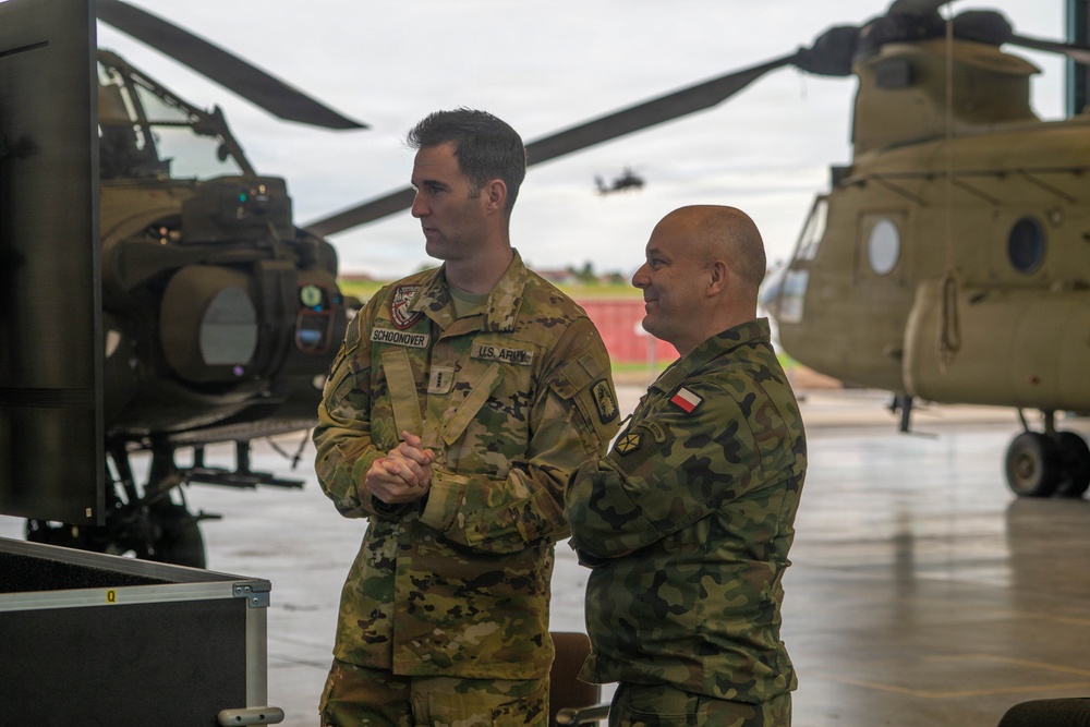 Two military personnel in different camouflage uniforms stand together on a helicopter flight line or tarmac. The taller individual wears a U.S. Army uniform with the name tape 'SCHOONOVER,' while the second individual wears a woodland camouflage pattern with what appears to be a Polish flag patch. They appear to be engaged in a briefing or discussion near military equipment, with CH-47 Chinook helicopters visible in the background.