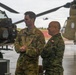 Two military personnel in different camouflage uniforms stand together on a helicopter flight line or tarmac. The taller individual wears a U.S. Army uniform with the name tape 'SCHOONOVER,' while the second individual wears a woodland camouflage pattern with what appears to be a Polish flag patch. They appear to be engaged in a briefing or discussion near military equipment, with CH-47 Chinook helicopters visible in the background.