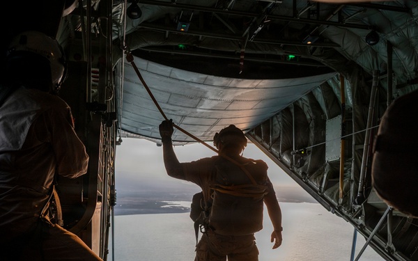 The image shows the interior of a military cargo aircraft with its rear ramp open in flight. Three personnel in military gear are visible in silhouette, with one standing at the edge of the open ramp looking out over a body of water below. The dramatic backlighting creates strong silhouettes against the sunlit water and sky visible through the open ramp.