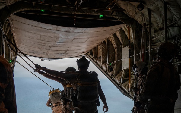 Military personnel are conducting a parachute jump operation from the open rear ramp of a large military cargo aircraft, likely a C-130 Hercules or similar transport, over a body of water. At least one jumper is already exiting the aircraft while others stand near the ramp edge. The image is taken from inside the aircraft looking toward the open ramp with ocean visible in the background.