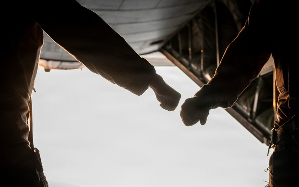 Two individuals in military-style clothing are performing a fist bump inside what appears to be the open rear cargo ramp of a military transport aircraft in flight. The image is backlit, rendering the figures largely in silhouette against a bright sky and distant terrain below. The composition emphasizes camaraderie and teamwork in an aerial military context.