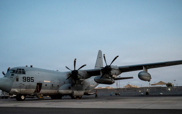 A military turboprop transport aircraft marked '985' is parked on a tarmac at dusk or dawn. The aircraft appears to be a C-130 Hercules variant in gray military livery, photographed at what appears to be a military air base. A navigation light is visibly illuminated on the wingtip, and ground support equipment is present nearby.
