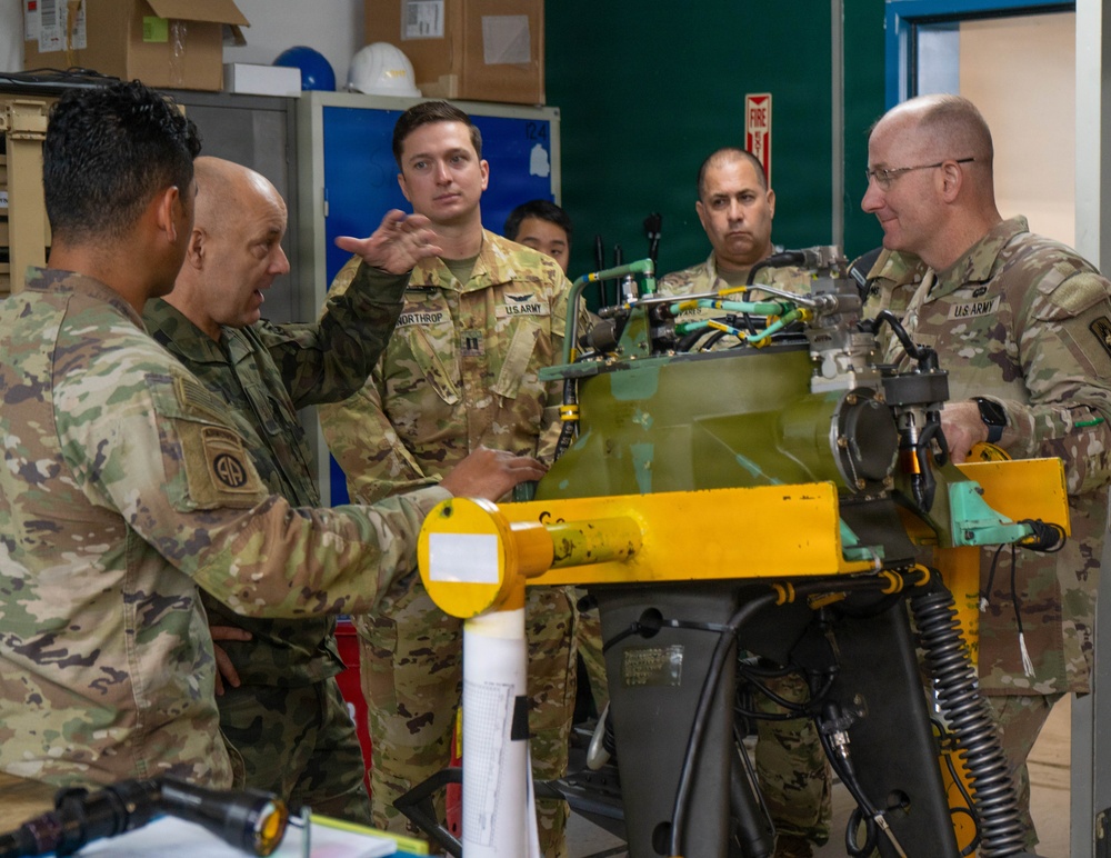 A group of U.S. Army soldiers in OCP (Operational Camouflage Pattern) uniforms are gathered around a piece of military equipment, likely an aircraft engine or mechanical component mounted on a yellow maintenance stand. The setting appears to be a maintenance facility or workshop, with one soldier gesturing and explaining something about the equipment to the others. The group includes personnel of varying ranks engaged in what appears to be a technical briefing or inspection.