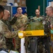 A group of U.S. Army soldiers in OCP (Operational Camouflage Pattern) uniforms are gathered around a piece of military equipment, likely an aircraft engine or mechanical component mounted on a yellow maintenance stand. The setting appears to be a maintenance facility or workshop, with one soldier gesturing and explaining something about the equipment to the others. The group includes personnel of varying ranks engaged in what appears to be a technical briefing or inspection.