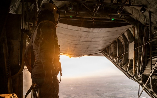 A military aircrew member stands at the open rear cargo ramp of a large military transport aircraft, likely a C-130 Hercules, looking out over a vast landscape below during what appears to be golden hour. The person is wearing flight gear and is connected to the aircraft via a safety cable or communication cord. The open ramp reveals a flat, arid terrain far below, suggesting the aircraft is at significant altitude.