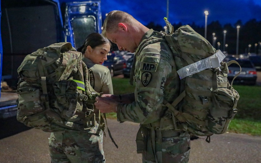 Two U.S. Army soldiers in Operational Camouflage Pattern (OCP) uniforms are adjusting or checking equipment in a parking lot during nighttime or early morning hours. The male soldier on the right wears a Military Police (MP) branch insignia patch on his left shoulder. Both soldiers are wearing large rucksacks and appear to be preparing for a ruck march or field exercise.