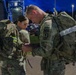 Two U.S. Army soldiers in Operational Camouflage Pattern (OCP) uniforms are adjusting or checking equipment in a parking lot during nighttime or early morning hours. The male soldier on the right wears a Military Police (MP) branch insignia patch on his left shoulder. Both soldiers are wearing large rucksacks and appear to be preparing for a ruck march or field exercise.