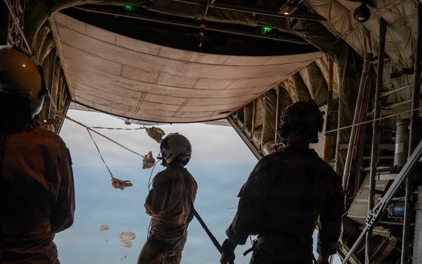 Three military personnel are standing at the open rear ramp of a large military cargo aircraft, likely a C-130 Hercules, observing an airdrop operation over a body of water. Multiple parachutes with cargo or supply bundles are visible descending toward the ocean below. The image is taken from inside the aircraft looking outward, with the crew members silhouetted against the exterior light.