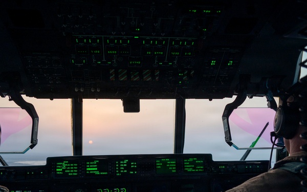 Two military pilots are seated in the cockpit of a large military aircraft, likely a C-130 Hercules or similar transport aircraft, flying at altitude during sunrise or sunset. The cockpit is illuminated by green-lit digital displays and instrument panels. A pink and purple sky with a visible sun is seen through the forward windscreen.