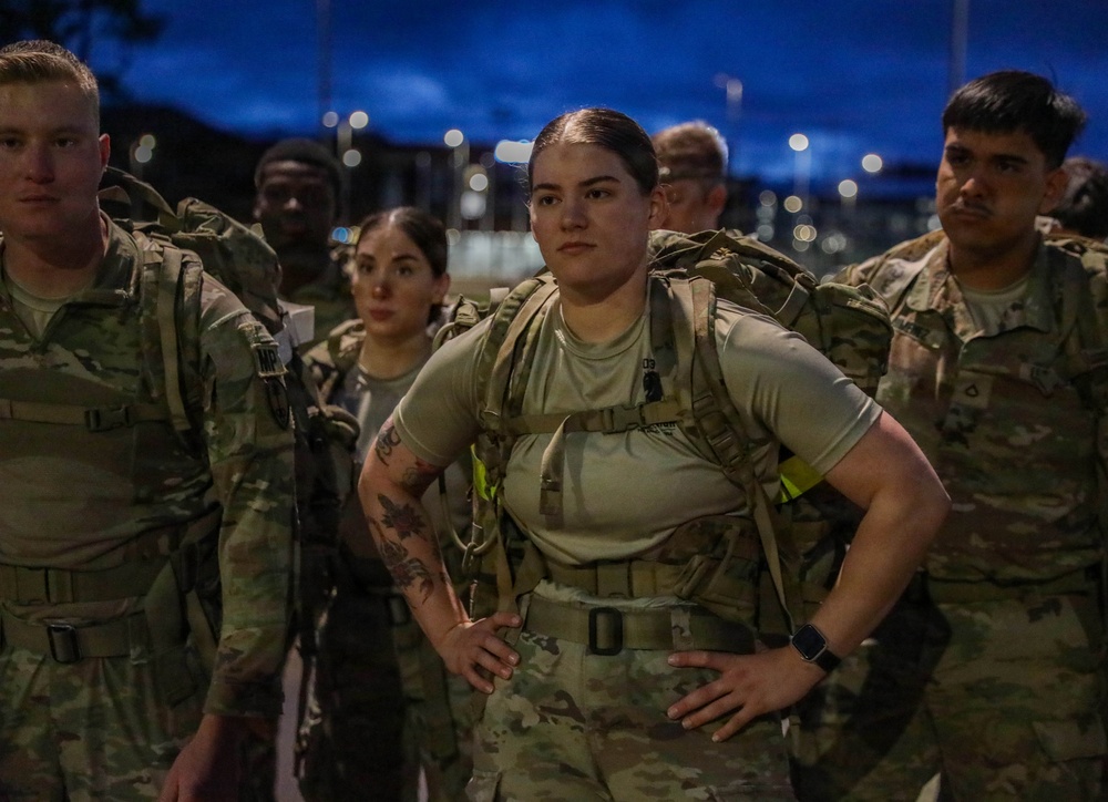 A group of military personnel in U.S. Army OCP (Operational Camouflage Pattern) uniforms are assembled outdoors during twilight or early morning hours. The central figure is a female soldier wearing a tactical backpack/ruck and standing with hands on hips in a confident posture. The setting appears to be a military installation or training area with artificial lighting visible in the background.