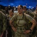A group of military personnel in U.S. Army OCP (Operational Camouflage Pattern) uniforms are assembled outdoors during twilight or early morning hours. The central figure is a female soldier wearing a tactical backpack/ruck and standing with hands on hips in a confident posture. The setting appears to be a military installation or training area with artificial lighting visible in the background.