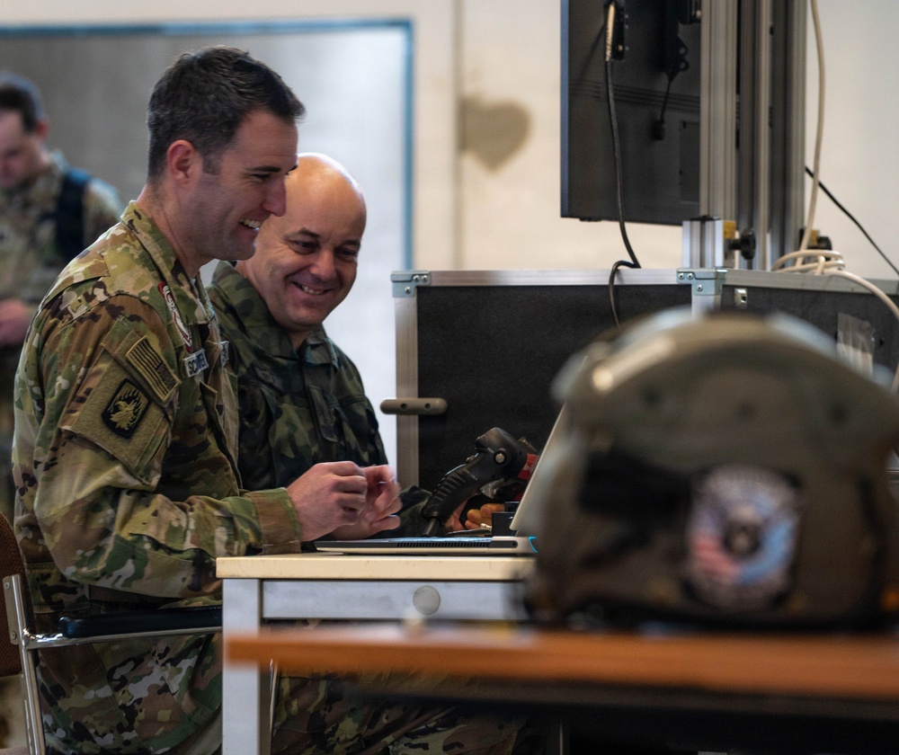 Two military personnel in different camouflage uniforms are seated at a workstation, smiling while examining equipment. The left individual wears US Army MultiCam/OCP uniform with a US flag patch and unit insignia, while the right individual wears a different camouflage pattern suggesting a foreign military. A monitor on a stand, a laptop, and what appears to be a military flight helmet are visible on and near the desk.