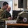 Polish General Piotr Trytek Strengthens Alliance During 12th CAB Visit Two military personnel in different camouflage uniforms are seated at a workstation, smiling while examining equipment. The left individual wears US Army MultiCam/OCP uniform with a US flag patch and unit insignia, while the right individual wears a different camouflage pattern suggesting a foreign military. A monitor on a stand, a laptop, and what appears to be a military flight helmet are visible on and near the desk.