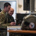 Two military personnel in different camouflage uniforms are seated at a workstation, smiling while examining equipment. The left individual wears US Army MultiCam/OCP uniform with a US flag patch and unit insignia, while the right individual wears a different camouflage pattern suggesting a foreign military. A monitor on a stand, a laptop, and what appears to be a military flight helmet are visible on and near the desk.