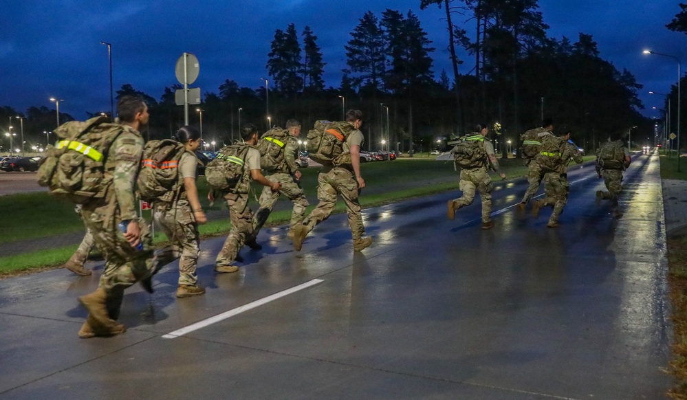 A group of military personnel in camouflage uniforms and full rucksacks are conducting a ruck march or physical training exercise on a wet road at dusk or dawn. The soldiers are wearing high-visibility reflective vests or bands over their gear, indicating a safety-conscious road movement. The setting appears to be a military installation or base, identifiable by the organized road infrastructure and surrounding facilities.