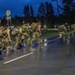 A group of military personnel in camouflage uniforms and full rucksacks are conducting a ruck march or physical training exercise on a wet road at dusk or dawn. The soldiers are wearing high-visibility reflective vests or bands over their gear, indicating a safety-conscious road movement. The setting appears to be a military installation or base, identifiable by the organized road infrastructure and surrounding facilities.