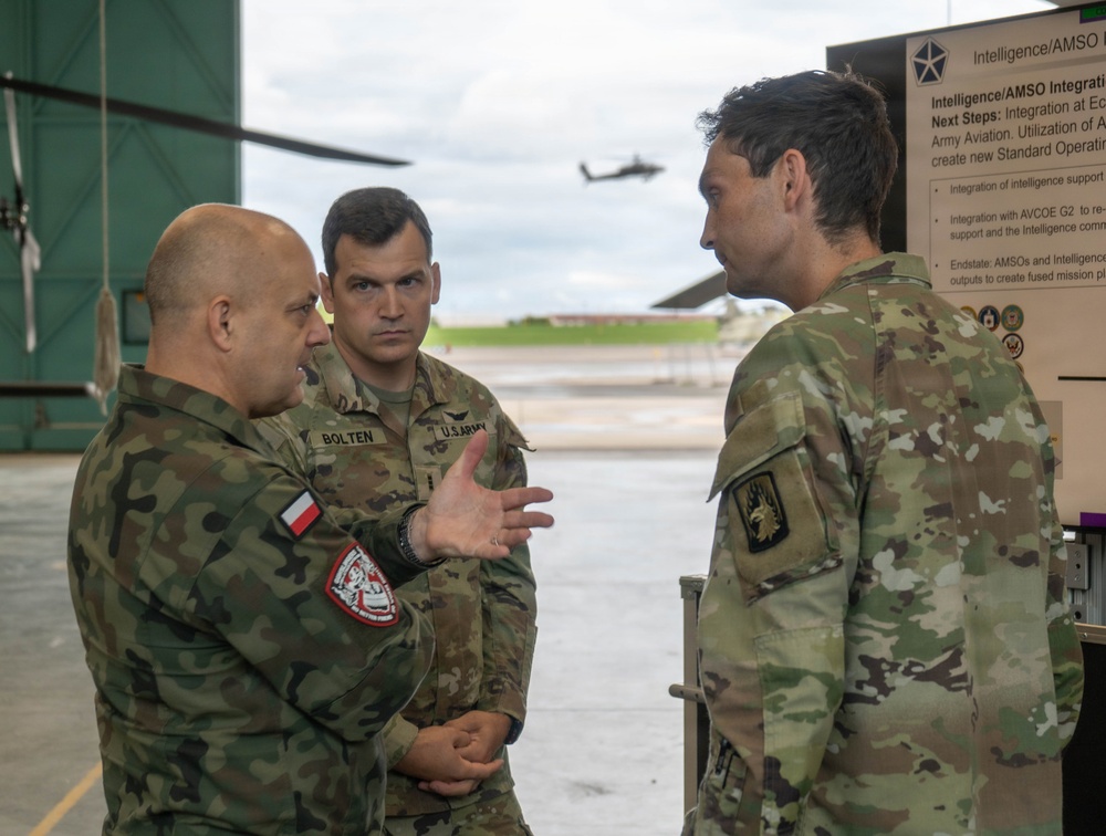 Three military personnel are engaged in a discussion near a briefing board labeled 'Intelligence/AMSO Integration' at what appears to be a military aviation facility. One individual wears a Polish military uniform with a Polish flag patch, while the other two wear U.S. Army OCP (Operational Camouflage Pattern) uniforms, one identified by a name tape reading 'BOLTEN'. A helicopter is visible in the background on the airfield.