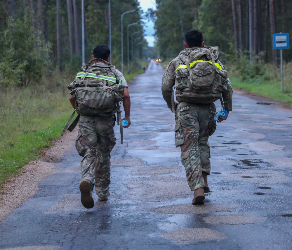Two soldiers in camouflage uniforms are moving along a wet, tree-lined road, viewed from behind. Both are carrying military rucksacks equipped with high-visibility yellow reflective strips, suggesting a ruck march or physical training exercise. The setting appears to be a forested military training area or rural road.