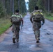 Two soldiers in camouflage uniforms are moving along a wet, tree-lined road, viewed from behind. Both are carrying military rucksacks equipped with high-visibility yellow reflective strips, suggesting a ruck march or physical training exercise. The setting appears to be a forested military training area or rural road.