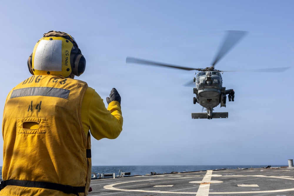 A naval flight deck crew member in a yellow safety vest and helmet is directing a military helicopter during a landing or departure operation on the flight deck of a U.S. Navy destroyer. The crew member's gear is marked 'DDG 53', identifying the vessel as USS John Paul Jones. The helicopter, likely an MH-60 Seahawk, hovers just above the helipad over open ocean.
