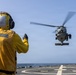 A naval flight deck crew member in a yellow safety vest and helmet is directing a military helicopter during a landing or departure operation on the flight deck of a U.S. Navy destroyer. The crew member's gear is marked 'DDG 53', identifying the vessel as USS John Paul Jones. The helicopter, likely an MH-60 Seahawk, hovers just above the helipad over open ocean.