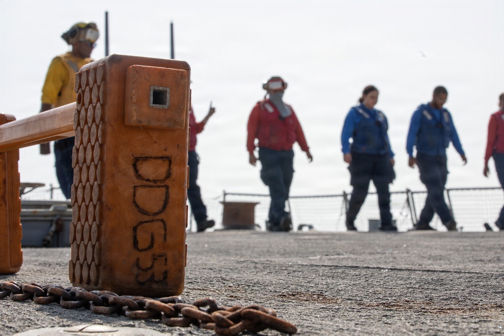 The image shows a naval vessel deck with crew members wearing color-coded jerseys and face coverings, indicating flight deck or operational duties. In the foreground, an orange chock or wheel chock is prominently marked with 'DDG53', identifying the ship as USS John Paul Jones (DDG-53). A heavy anchor chain lies on the deck surface near the chock.