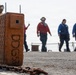 The image shows a naval vessel deck with crew members wearing color-coded jerseys and face coverings, indicating flight deck or operational duties. In the foreground, an orange chock or wheel chock is prominently marked with 'DDG53', identifying the ship as USS John Paul Jones (DDG-53). A heavy anchor chain lies on the deck surface near the chock.
