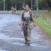 A female soldier in U.S. Army OCP (Operational Camouflage Pattern) uniform is conducting a ruck march along a wet paved road, carrying a large military rucksack. She is wearing a unit t-shirt over or instead of her uniform top, with camouflage trousers and tan combat boots. Additional soldiers are visible in the background performing the same activity.