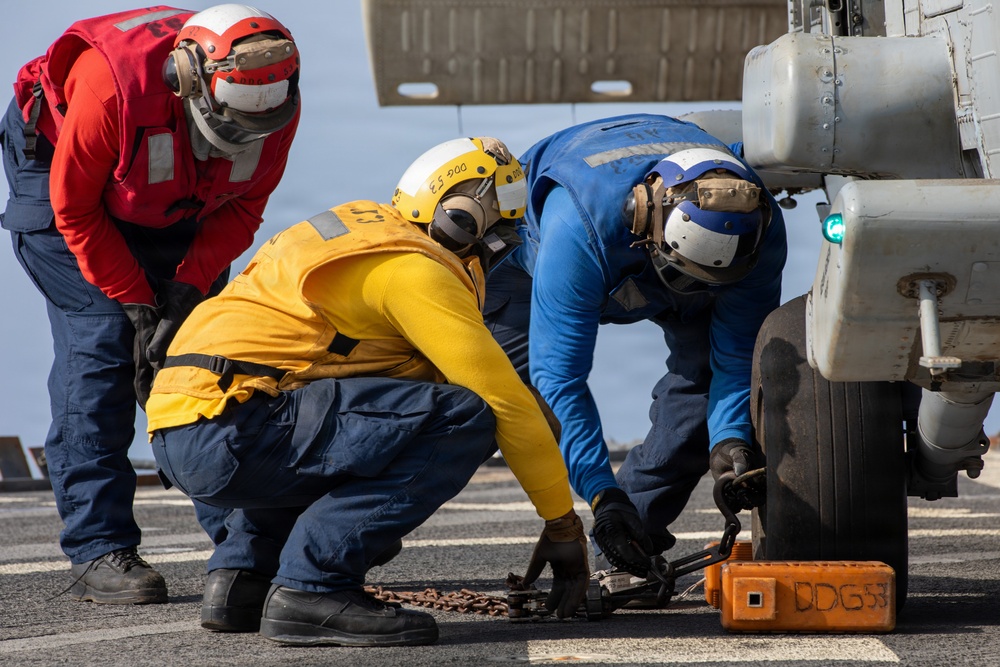 Three naval personnel are working on an aircraft landing gear on what appears to be a ship's flight deck. They are securing or chocking the wheel of an aircraft, with one orange chock block visible marked 'DDG 53'. The crew members are wearing color-coded flight deck jerseys and helmets indicating their respective roles.