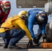 Three naval personnel are working on an aircraft landing gear on what appears to be a ship's flight deck. They are securing or chocking the wheel of an aircraft, with one orange chock block visible marked 'DDG 53'. The crew members are wearing color-coded flight deck jerseys and helmets indicating their respective roles.