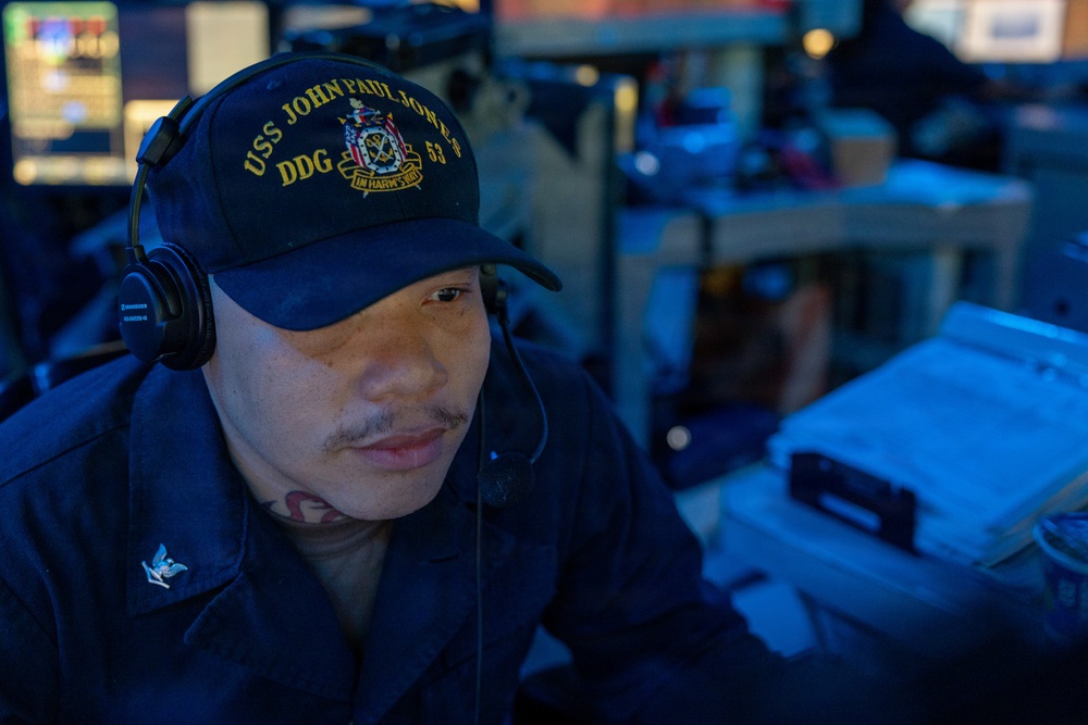 A U.S. Navy sailor is seated at a workstation aboard the USS John Paul Jones (DDG-53), wearing a ship's ball cap and headset. The image is taken in a dimly lit combat information center or operations room with blue ambient lighting. The sailor appears focused on tasks at the workstation.