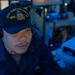 A U.S. Navy sailor is seated at a workstation aboard the USS John Paul Jones (DDG-53), wearing a ship's ball cap and headset. The image is taken in a dimly lit combat information center or operations room with blue ambient lighting. The sailor appears focused on tasks at the workstation.