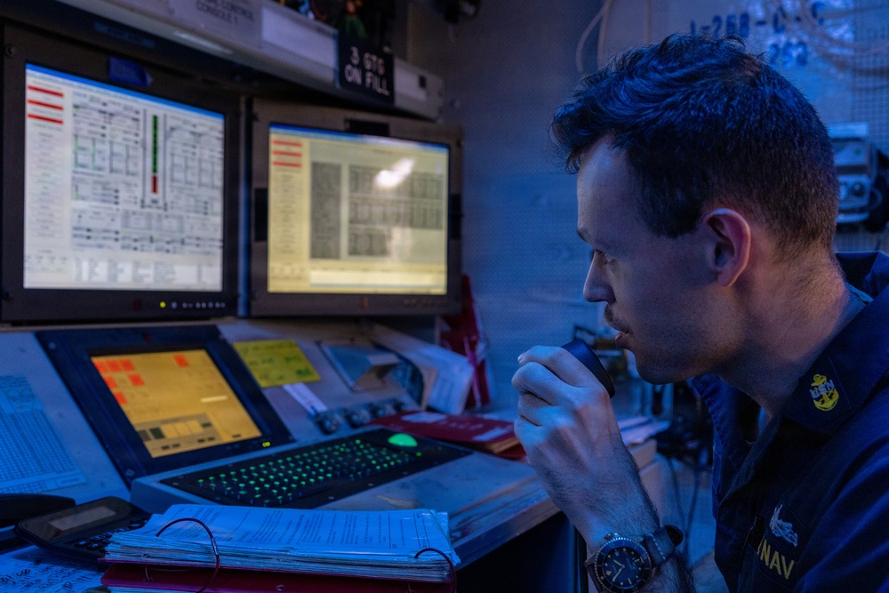 A U.S. Navy sailor is seated at a multi-monitor control console in a dimly lit operations room, holding a communication handset to his mouth. The environment appears to be aboard a naval vessel, indicated by the 'NAVY' text on his uniform and the specialized monitoring equipment surrounding him. The blue ambient lighting and multiple status-display screens suggest an active watch station or engineering control room.