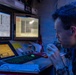 A U.S. Navy sailor is seated at a multi-monitor control console in a dimly lit operations room, holding a communication handset to his mouth. The environment appears to be aboard a naval vessel, indicated by the 'NAVY' text on his uniform and the specialized monitoring equipment surrounding him. The blue ambient lighting and multiple status-display screens suggest an active watch station or engineering control room.