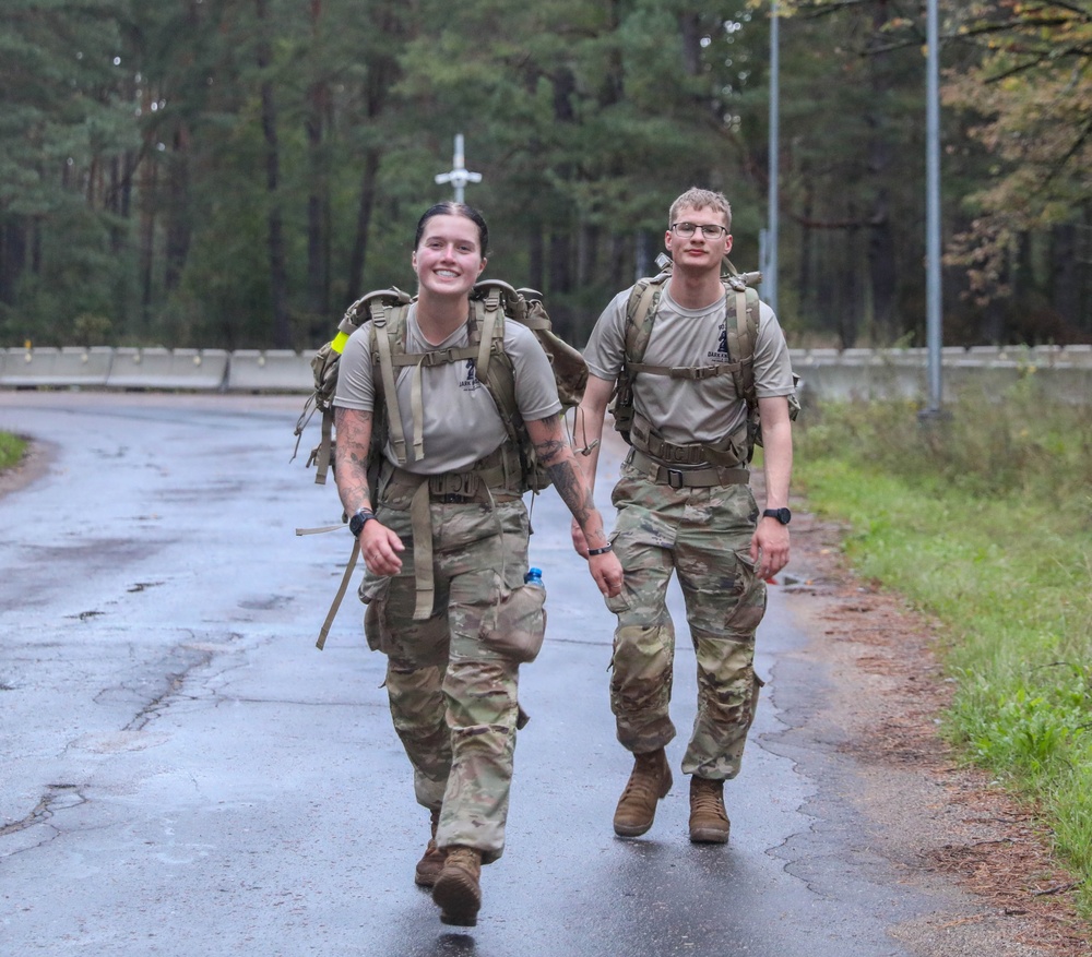 Two military personnel in camouflage uniforms and tactical gear are walking along a wet road through a forested area. Both are carrying large rucksacks and wearing matching unit t-shirts under their gear, suggesting participation in a ruck march or physical training event. The soldier in front is smiling broadly, indicating a positive or accomplished mood.
